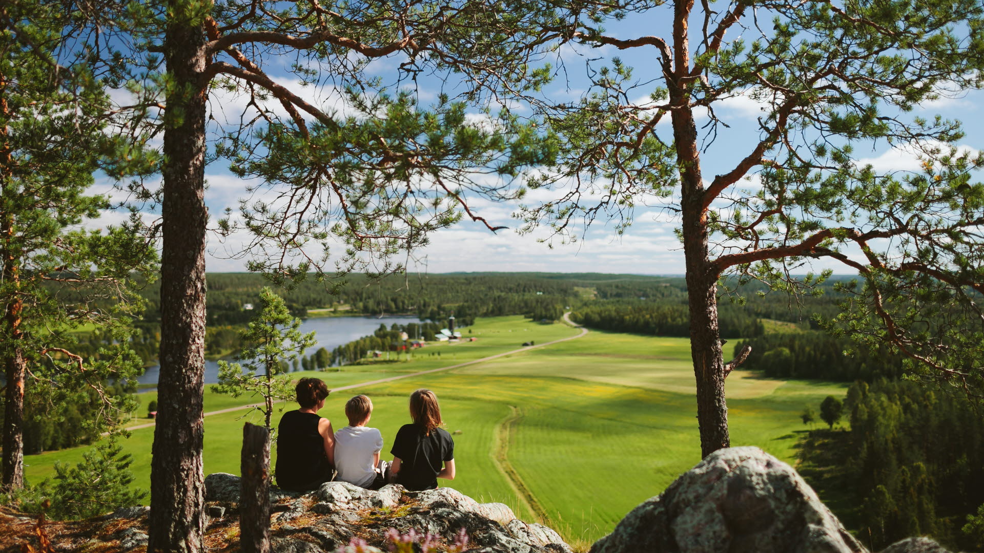 Tre personer sitter på en höjd (Falkberget) och ser ut över ett odlingslandskap med en sjö